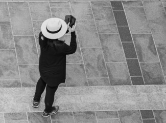 Tourist taking a photo on a street of a city with her smartphone top view in monochrome. Woman in a hat taking pictures.
