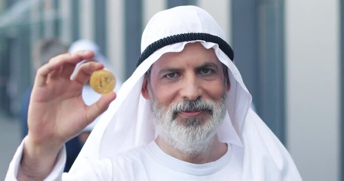Portrait of smiling arab sheikh in national clothes holding bitcoin and showing it to the camera. Senior male standing at the street, smiling and looking at camera. Selling and business concept