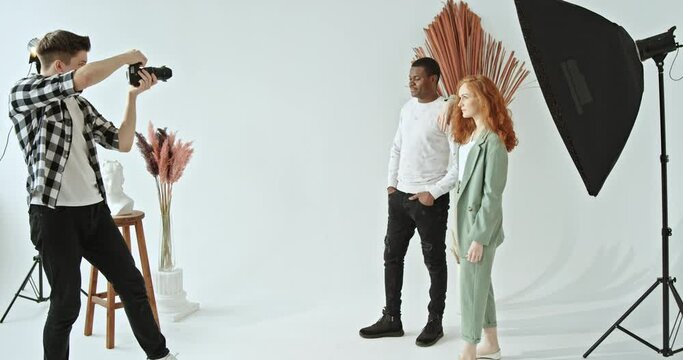 Process photoshoot multiracial couple dark-skinned guy and red-haired girl standing together in a bright photo studio posing in front of a photographer