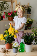 a little blonde girl with a headband on her head and wearing work gloves sprays or waters indoor flowers. Concept of spring time, home gardening, child house-help. Lifestyle, space for text. 