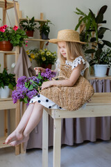 a little blonde girl in a dress with black polka dots and a straw hat with a bouquet of lilac flowers is sitting on a wooden chair in the room. lifestyle, high quality photo