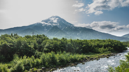 The picturesque Vilyuchinsky volcano with snow-covered slopes against the background of blue sky and clouds.  At the foot there is green vegetation, the river flows along a rocky bed. Kamchatka.