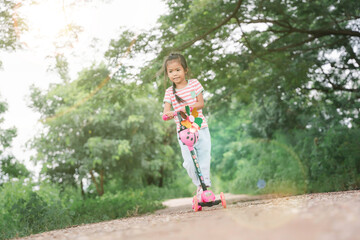 Asia children learn to ride scooters in a park on a summer day. Preschooler girl riding a roller. Kids play outdoors with scooters. Active leisure and outdoor sport for children