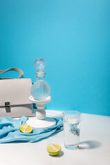 Fashionable bag of light nude color against a blue wall with a decanter of water, lime fruit and a glass with liquid on a white table in bright sunlight 