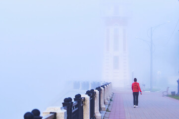 Fog on the city embankment. Lighthouse in strong blur. A girl in a red sports jacket at morning...
