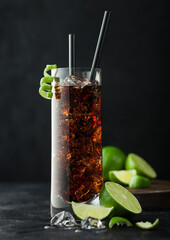 Cuba Libre cocktail in highball glass with ice and lime peel with straw and fresh limes on black background.