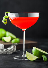Cosmopolitan cocktail in luxury crystal glass with lime peel and fresh limes with strainer on black background.