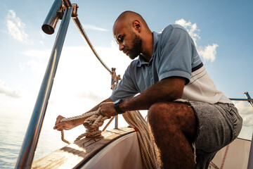 Young african american sailor tying ropes on sailboat in the sea on sunset - obrazy, fototapety, plakaty