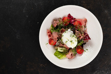 A beautiful Fresh salad with meat roast beef vegetables lettuce leaves poached egg cucumbers bell pepper olives red onions is decorated on a white plate on a black background large serving Restaurant