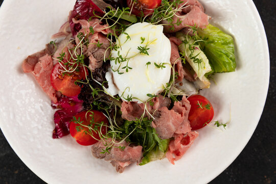 A beautiful Fresh salad with meat roast beef vegetables lettuce leaves poached egg cucumbers bell pepper olives red onions is decorated on a white plate on a black background large serving Restaurant