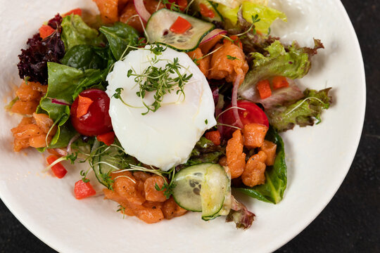 A beautiful fresh salad with salmon red fish vegetables lettuce leaves poached egg cucumbers bell peppers olives red onions is decorated on a white plate on a black background close up
