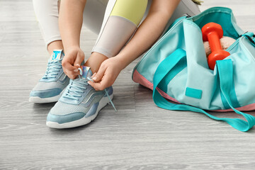 Woman tying shoelaces and bag with dumbbells on light wooden floor