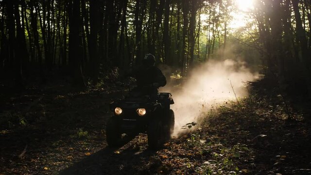 An quadbike rider rides at high speed through a dark forest. Behind him the sun shines illuminating his trail with pollen.