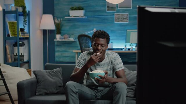 Black adult watching comedy movie on television while eating popcorn from bowl. African american person laughing and looking at TV with snack. Man of african ethnicity having fun