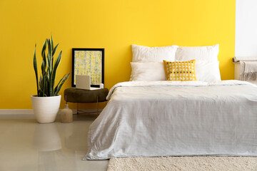 Bedroom with comfortable bed, laptop on pouf and houseplant near color wall
