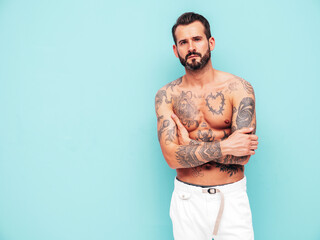 Fototapeta premium Portrait of handsome confident stylish hipster lambersexual model. Sexy modern man. Naked torso with tattoos.Fashion male posing in studio near light blue wall. Crossed arms