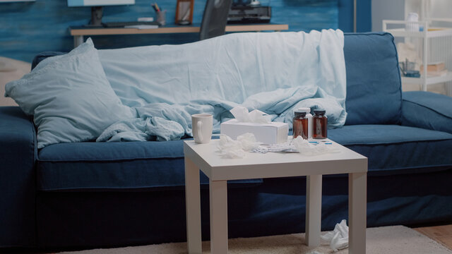 Empty living room with healthcare treatment on table for sick person. Nobody in place with medicine, bottles of pills, capsule tablets, tea and tissues used by adult with disease.