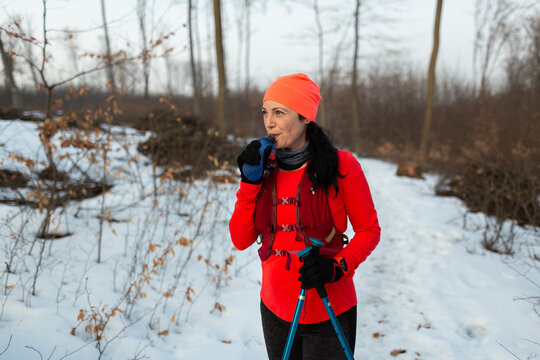 Female Runner Drinking Water from Soft Flask in Woods on Winter Day. Woman Wearing Hydration Vest Taking Break From Trail Running in Woods and Sipping Drink from Sports Bottle On Cold Winter Day.