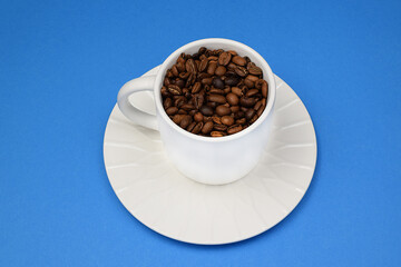 White cup and saucer filled with roasted coffee beans on a blue background. Side and top view.