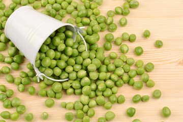 Green peas are poured out of a small white bucket on a wooden background.The concept of growing and caring for agriculture,biology,agronomy,breeding,vegetarianism,protein production,sports nutrition.