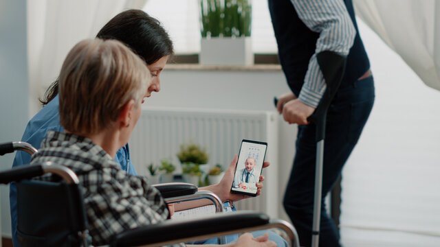 Senior Patient Talking To Doctor On Video Call In Nursing Home. Old Woman With Chronic Disability In Wheelchair Using Telemedicine And Online Conference While Nurse Holding Smartphone