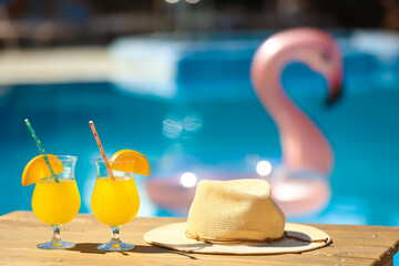 Glasses of cocktails and hat on table near swimming pool