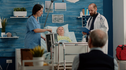 Obraz premium Doctor and nurse doing checkup visit for ill patient in hospital bed. Medical team consulting retired woman with sickness while having bottle of pills and equipment for recovery.