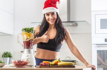 Thumbs up by a woman in Santa's hat standing in workout clothes next to healthy smoothie maker full of healthy stuff