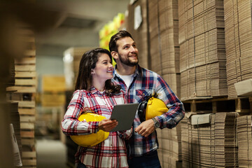 Warehouse overview. A man and a woman in plaid shirts with yellow protective helmets under their arms inspect and check the condition of the raw material warehouse. They check on a digital tablet