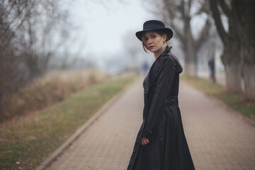 A middle-aged woman in a black raincoat and hat alone in the park. Autumn mood of November. Gloomy rainy foggy weather. Loneliness bad mood depression. Despair. Halloween time. Mental health lifestyle