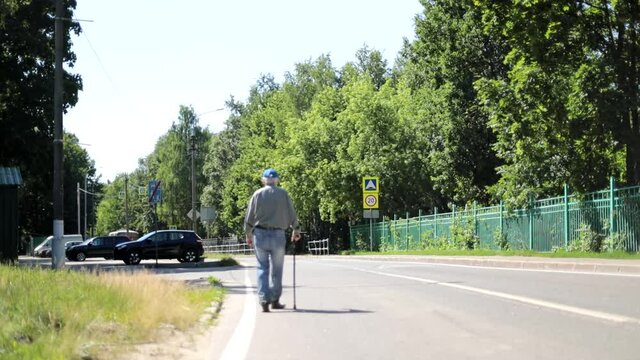 An old man walks down the street. An elderly man with a walking stick walks through a village in Russia. A Russian pensioner walks down the road.