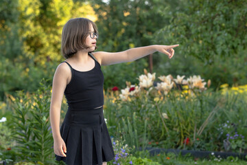 Girl in glasses in black clothes points with her hand at something in a blooming garden