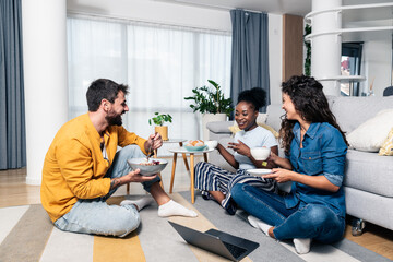 Three young carefree friends two women and one man sitting in the apartment on the floor listening to music on a laptop eating and having fun for a free weekend without worries