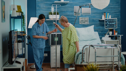 Sick old woman walking with IV drip bag and nurse giving assistance in nursing home. Medical...