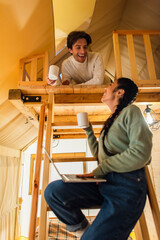 Low angle view of smiling interracial couple with cups and laptop looking at each other in glamping house