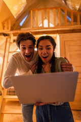 Amazed man hugging asian girlfriend with laptop in glamping house