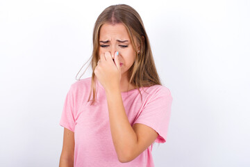 beautiful blonde girl wearing pink t-shirt on white background, holding his nose because of a bad...