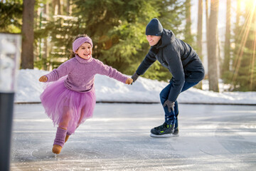 a little girl in a pink sweater and a fluffy skirt on a sunny winter day rides with her dad on an...