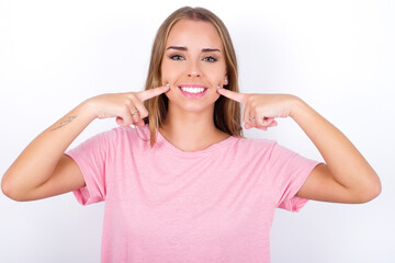 Strong healthy straight white teeth. Close up portrait of happy beautiful blonde girl wearing pink t-shirt on white background with beaming smile pointing on perfect clear white teeth.