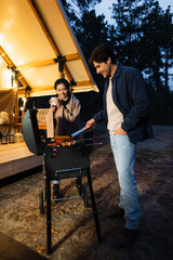 Fototapeta na wymiar Smiling interracial couple with cup cooking on grill on glamping terrace