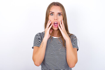 Speechless beautiful blonde girl wearing striped t-shirt on white background keeps hands near opened mouth reacts to shocking news stares wondered at camera