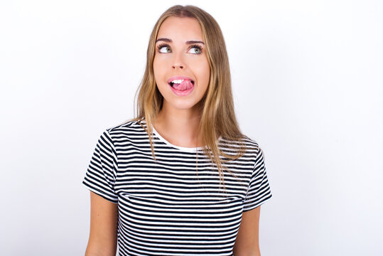 Funny beautiful blonde girl wearing striped t-shirt on white background makes grimace and crosses eyes plays fool has fun alone sticks out tongue.