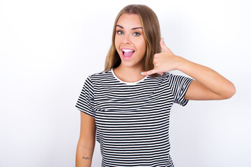 Fototapeta premium beautiful blonde girl wearing striped t-shirt on white background makes phone gesture, says call me back again, has glad expression.
