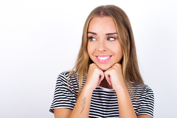 beautiful blonde girl wearing striped t-shirt on white background holds hands under chin, glad to hear heartwarming words from stranger