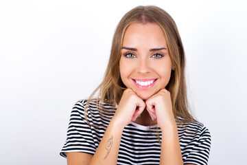 Fototapeta premium Satisfied beautiful blonde girl wearing striped t-shirt on white background touches chin with both hands, smiles pleasantly, rejoices good day with lover