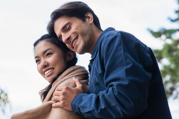 Low angle view of man with closed eyes hugging asian girlfriend in blanket outdoors