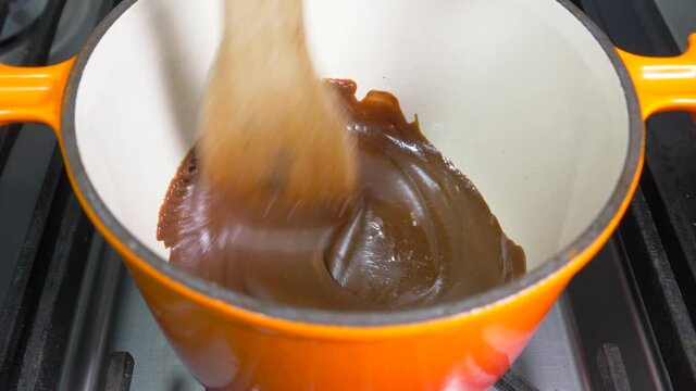 Closeup POV Shot Of A Wooden Spatula Being Used To Stir Warm, Thick, Melted Brown Chocolate In An Orange, Enamel Coated Pan, Over A Gentle Heat On A Cooker Hob.