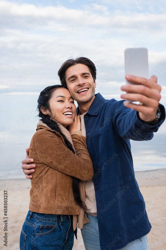 Wall mural smiling interracial couple hugging and taking selfie on blurred smartphone on beach