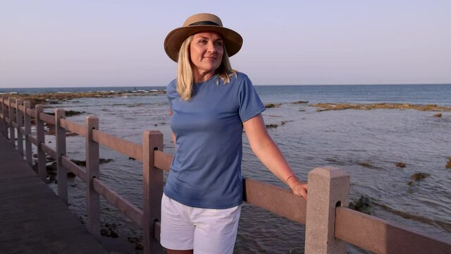 Smiling middle aged woman wearing blue t-shirt and straw hat and standing at sunset on the wooden bridge with hands on it. T-shirt mockup