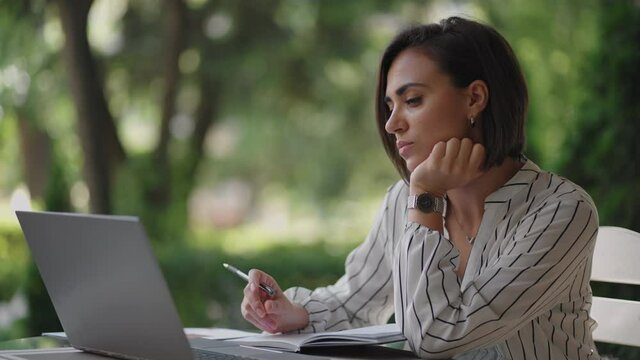 Student Woman Brunette arabic Hispanic ethnos studying remotely via the Internet while sitting in a summer cafe with a laptop and writing down a pen and a notebook while taking notes on the webinar